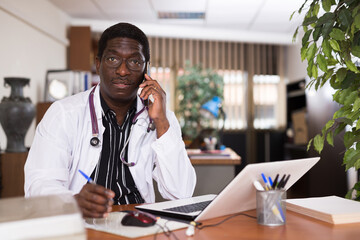African american doctor talking with patient on mobile phone, sitting at workplace in clinic, taking notes