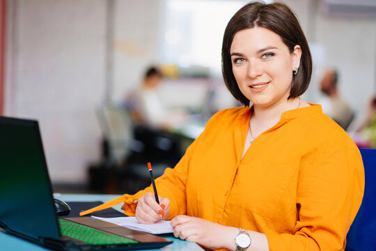 a woman wearing a orange blouse with laptop in a office. 