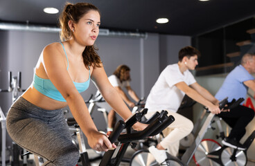 Fototapeta na wymiar Concentrated young fit girl working out on stationary bicycle in gym. Healthy active lifestyle concept