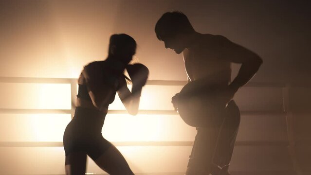 Sporty couple building muscle strength and burning calories in the gym. Close-up view of aggressive woman in boxing gloves punching her coach during a tough workout. High quality 4k footage