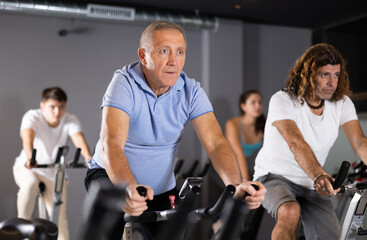 Fototapeta na wymiar Positive mature male riding exercise bike during cycling class in modern gym