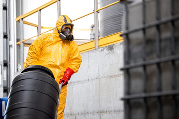 Worker in protective hazmat suit, gas mask, gloves and boots moving barrels with chemicals on...