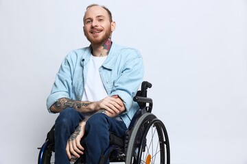 A man in a wheelchair smile, copy space, with tattoos on his arms sits on a gray studio background,...