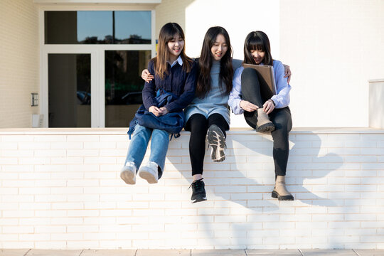 Group of Asian girls outdoor students. The 3 women are sitting on the wall while watching one of them tie her bootlaces. Education, friendship and concept of people.