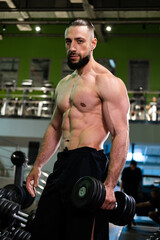 Fototapeta na wymiar A handsome muscular male bodybuilder with a beard with a pumped-up torso holds dumbbells in the gym