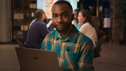 African american smiling employee working on laptop portrait, slow motion, front view. Office worker holding portable computer, looking at camera medium shot, business meeting in background