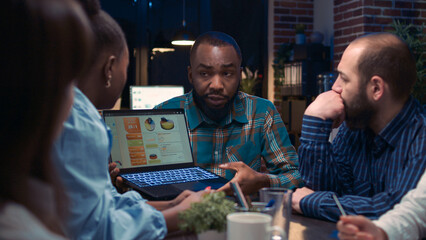 Fototapeta na wymiar Diverse coworkers brainstorming, african american employee showing data charts on laptop screen. People planning strategy in business meeting, discussing presentation, team communication