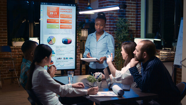 African american businesswoman speaking in business meeting presentation, holding report clipboard. Diverse team discussing revenue statistics, planning sales strategy, looking at graphs on screen