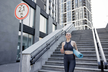 Pretty fitness trainer carrying yoga mat, while walking down stairs outdoors.