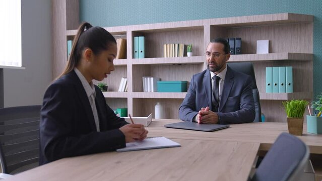 A man in a suit, the boss in the office sits at the desk, talks to a young woman, they discuss work issues, the worker is upset, the boss is dissatisfied with the work of the subordinate