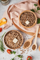 Wooden bowls of tasty buckwheat porridge with butter and vegetables on grey table