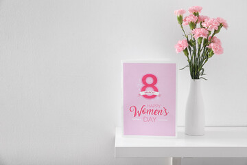 Vase with carnation flowers and greeting card for Women's Day on shelf in light room