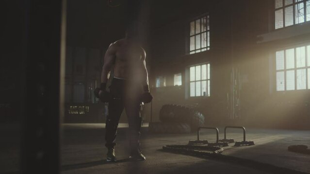 Slow-motion shot of a man doing dumbbell workout for triceps at sports gym. Tyres and gym equipments in the background