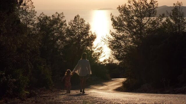 couple moves away from the camera. Mom and little daughter walk hand in hand down slope at sunset. Traveling around world together. mother holds her little child on steep descent from mountain.