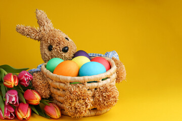Straw bunny basket with easter colorful eggs und fresh tulips on yellow background. Happy Easter card. Close up