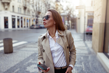 Fototapeta premium Fashionable woman in sunglasses is standing with phone on city street background and looking at side