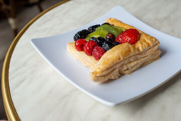 A square fruit filled French puff pastry on a white square plate. The cream and custard filled dessert have strawberries, kiwi, and blackberries on top with a sugar glaze. The pastry is flaky.	