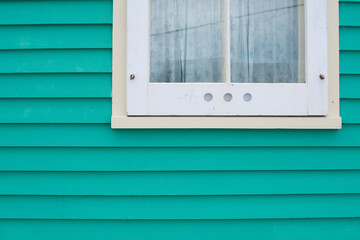 A small single casement window in a white narrow wood beveled clapboard siding exterior wall of a vintage house. The glass window has six panes with green decorative trim on the outside edge. 