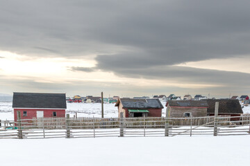 Multiple small red wooden barns and pens for storing farm animals. There are multiple pens with log fencing in the field. The sky is grey and stormy and the ground is covered in fresh white snow.