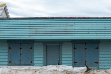 A blue color exterior wooden wall of a vintage warehouse or garage with multiple double doors and a single wooden door in the middle. The wooden doors are a darker shade of blue. It's winter with snow