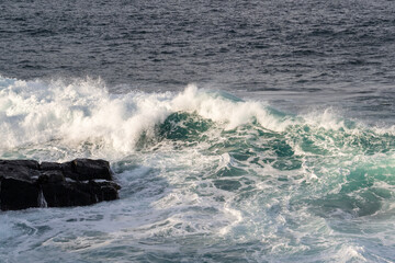 A seascape of large waves hitting rocks along the shore.  The waves are breaking and sending a spray back into the ocean. The water is a deep blue and the waves are made of bright white foam.