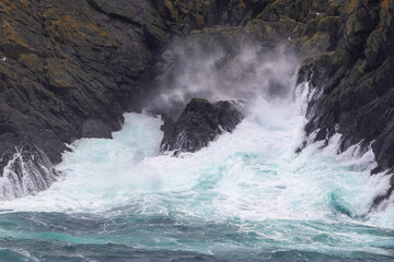 A seascape of a large waves hitting rocks along the shore.  The waves are breaking and sending a spray back into the ocean. The water is a deep blue and the waves are made of a bright white foam.
