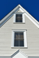 An upward view of a dark blue sky and an exterior gable end of a white wooden vintage house with two small double hung windows. The wall has narrow clapboard siding and wide moldings.  