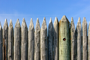 A row of old gray weathered log stakes lined to make a seawall or fence. The wall of logs has a pointy edge post with a sharp tip. The background is daytime outdoors of blue sky with white clouds.