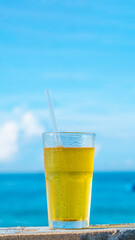 Big glass drink plastic straw green tea looks like yellow beer or kombucha blue sea white clouds background water drops condensed