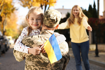 Daughter hugging her father in military uniform and holding Ukrainian flag on city street. Family...