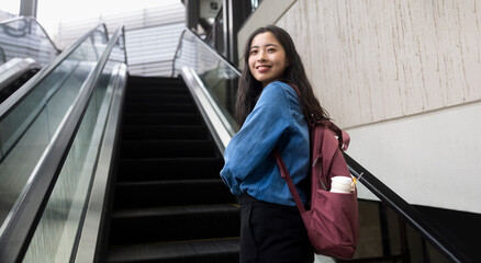 Portrait of a young Asian girl in a jeans jacket riding the escalator of a shopping mall with a backpack 