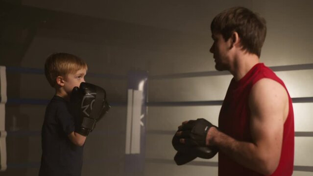 Close-up of young and adult sparring partners looking focused and motivated. Portrait of a skilled boxer teaching little boy to fight in boxing gloves. High quality 4k footage