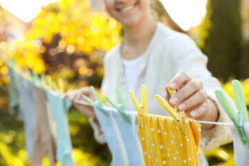Closeup view of smiling woman hanging baby clothes with clothespins on washing line for drying in...