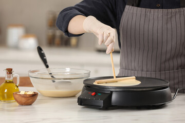 Fototapeta na wymiar Woman cooking delicious crepe on electric pancake maker at white marble table in kitchen, closeup