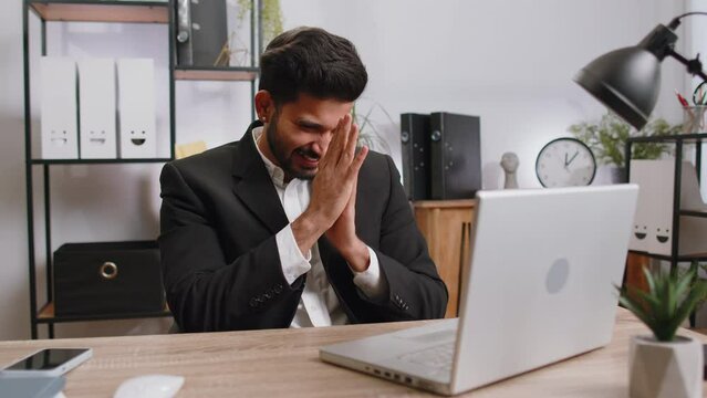Sneaky cunning bearded indian businessman with tricky face gesticulating and scheming evil plan, thinking over devious villain idea, cunning cheats, jokes and pranks at office workplace with laptop