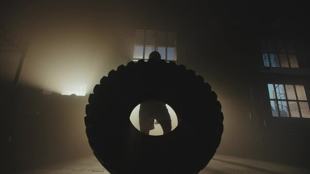 Muscular young shirtless man exercising with giant tire during training in gym, front view 