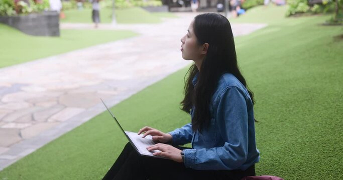 Portrait of a cute asian student girl coding IT laptop outdoors on grass in an open mall outside