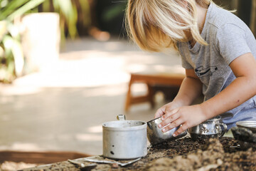 Little blonde preschool boy playing in outdoor mud kitchen with pots and pans at preschool
