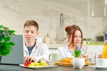 Happy childrens, kids having healthy breakfast at home and using digital devices. kids playing and eating. Using notebook in kitchen at breakfast time