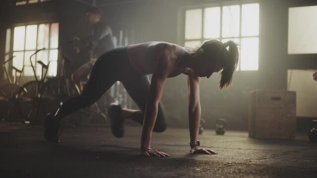 Attractive young woman wearing sports bra doing pushups at a gym while two other men exercising in the background. Slow motion handheld medium shot