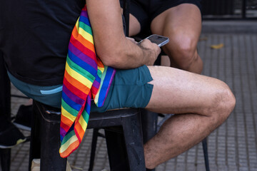 Gay couple with Rainbow Flag sitting outside bar chatting on smartphone at Pride Event. LGBTQ and diversity concept. Copy space. 