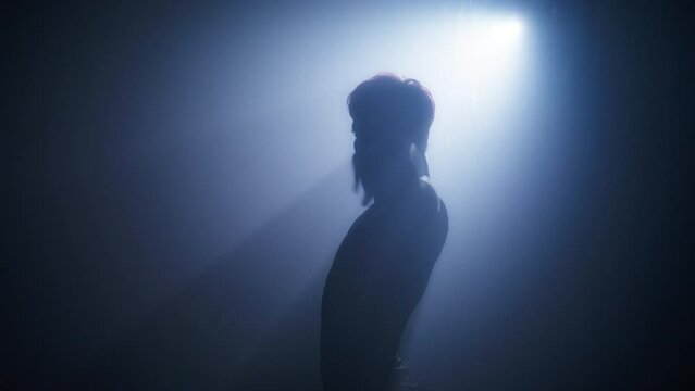 Seductive woman silhouette dancing in dark room with smoke and bright spotlight on background. Slim athletic body woman dancing, touching her neck and body in a dark room with backlit RED shot 4K