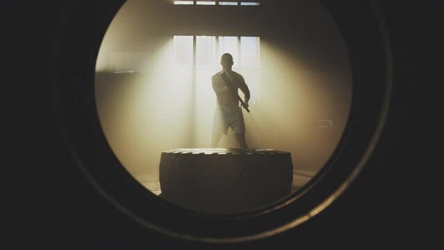View through the tyre hole of a shirtless handsome man hitting large tractor tyre with a hammer in slow-motion at gym