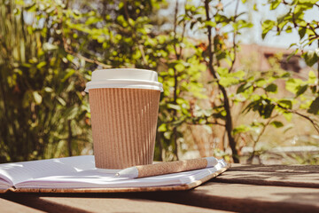 Eco recycling paper cup with coffee or tea on kraft paper with empty paper notebook on wooden bench. Concept of study work outdoors. Take away coffee to go. Copy space for text. Disposable Cardboard