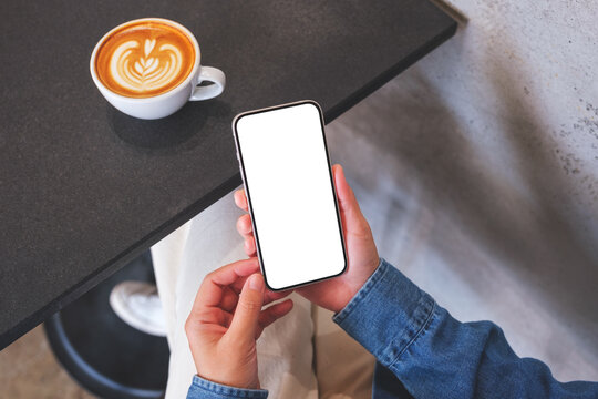 Top view mockup image of a woman holding mobile phone with blank desktop screen with coffee cup on the table
