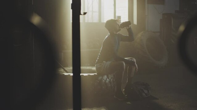 Slow-motion shot of a young man sitting in the gym after exercise and drinking water from plastic bottle 