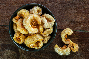 Top view of homemade crispy delicious deep fried pork rinds in black ceramic bowl on wooden background.