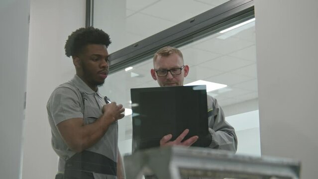 Two multi cultural installers in uniform sets up security cameras in modern college using laptop. They talk, check CCTV cameras in computer program. Concept of installation and surveillance system.