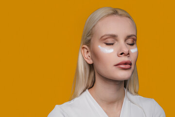 Caucasian blonde woman with cream on her cheek, enjoying skin care, posing with closed eyes isolated over yellow background. Beauty, tenderness concept.