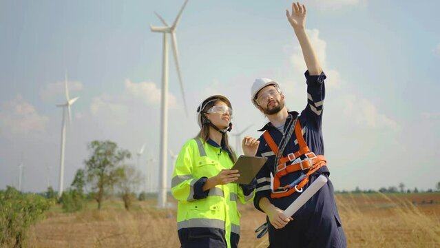 Caucasian engineer man discuss together with co-worker woman hold tablet and blueprint stay in front of row of windmill or wind turbine, wind turbine generate electricity, Environmentally friendly